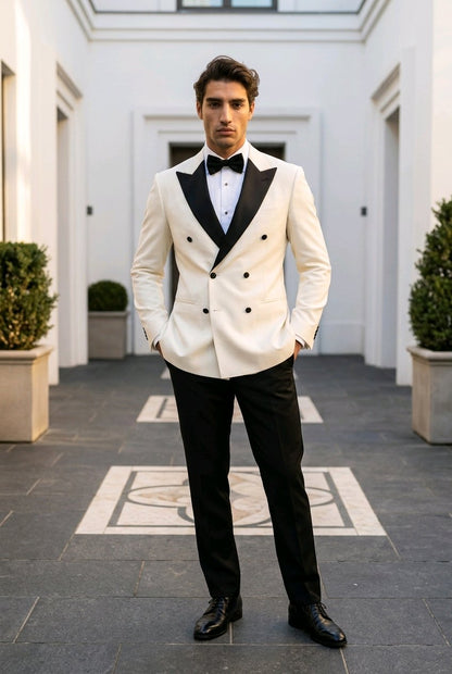 A man wearing a double-breasted tuxedo jacket in off-white with black satin lapels, paired with black trousers, a black bow tie, and black dress shoes, standing in a grand courtyard.