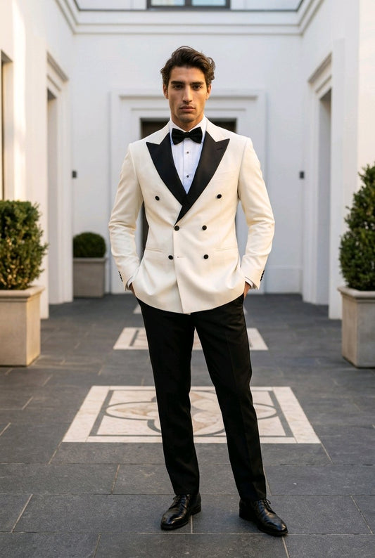 A man wearing a double-breasted tuxedo jacket in off-white with black satin lapels, paired with black trousers, a black bow tie, and black dress shoes, standing in a grand courtyard.