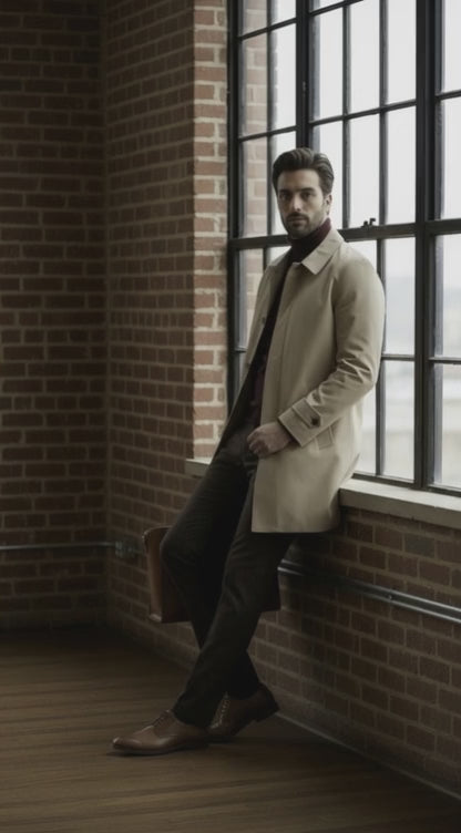 Man in a beige coat leaning against a brick wall with large windows.