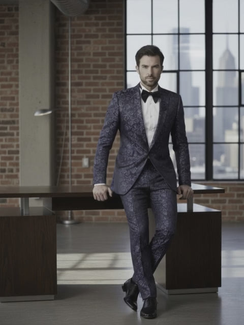 Man wearing a dark patterned suit with a white shirt and black bow tie in an urban setting.